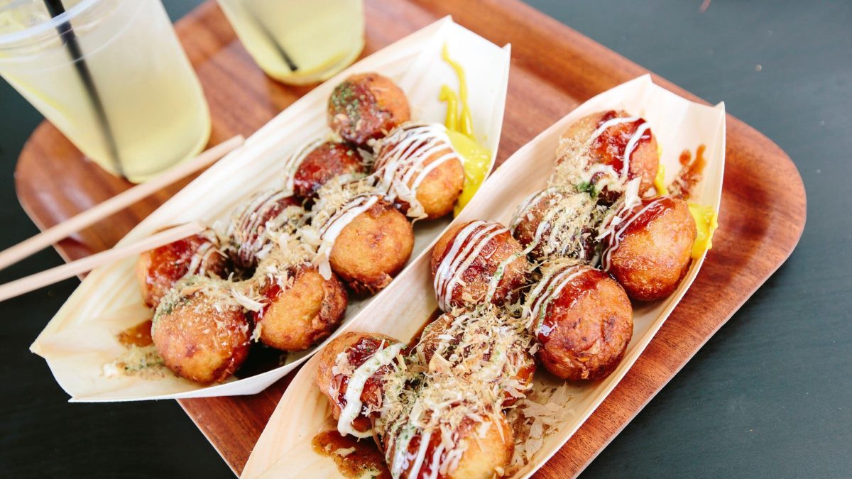 Takoyaki in zwei Papierschiffchen auf einem Holzbrett.