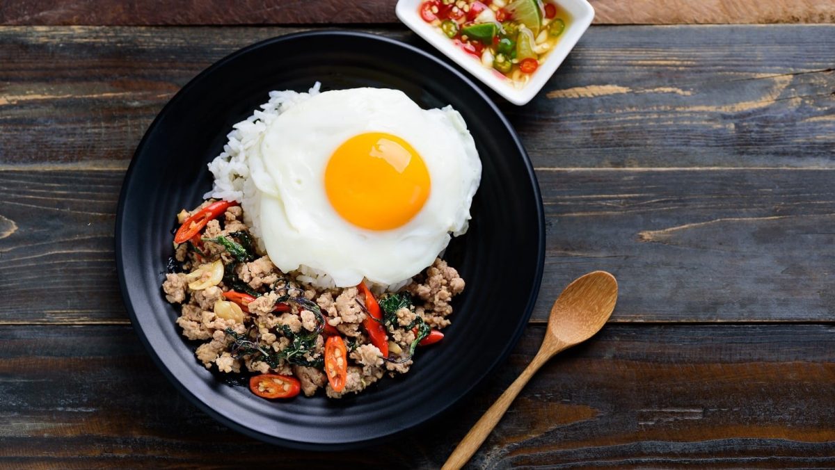 Pad Krapao getoppt von einem Spiegelei in einem schwarzen Teller serviert neben einem Holzlöffel und Chilidip