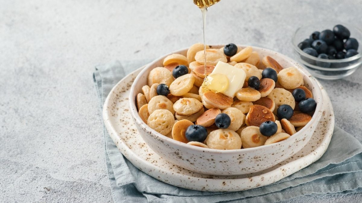 Weiße Schale mit kleinen Pancakes und Blaubeeren auf grauem Untergrund in seitlicher Aufnahme.
