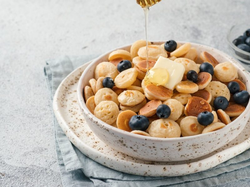 Weiße Schale mit kleinen Pancakes und Blaubeeren auf grauem Untergrund in seitlicher Aufnahme.