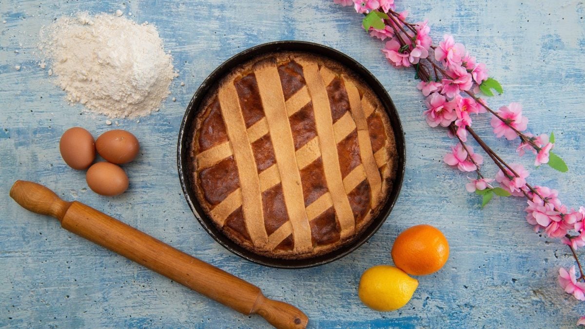 Italienische Pastiera napoletana italienisches Osterbrot in schwarzer Backform auf blauem Untergrund. Daneben Zweig mit rosa Orangenblüten, Mehl, drei Eier, Nudelholz sowie je eine Orange und Zitrone. Aufnahme aus der Vogelperspektive.