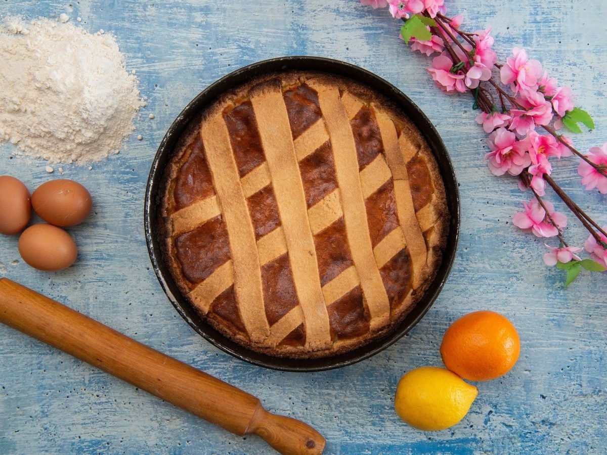 Italienische Pastiera napoletana italienisches Osterbrot in schwarzer Backform auf blauem Untergrund. Daneben Zweig mit rosa Orangenblüten, Mehl, drei Eier, Nudelholz sowie je eine Orange und Zitrone. Aufnahme aus der Vogelperspektive.