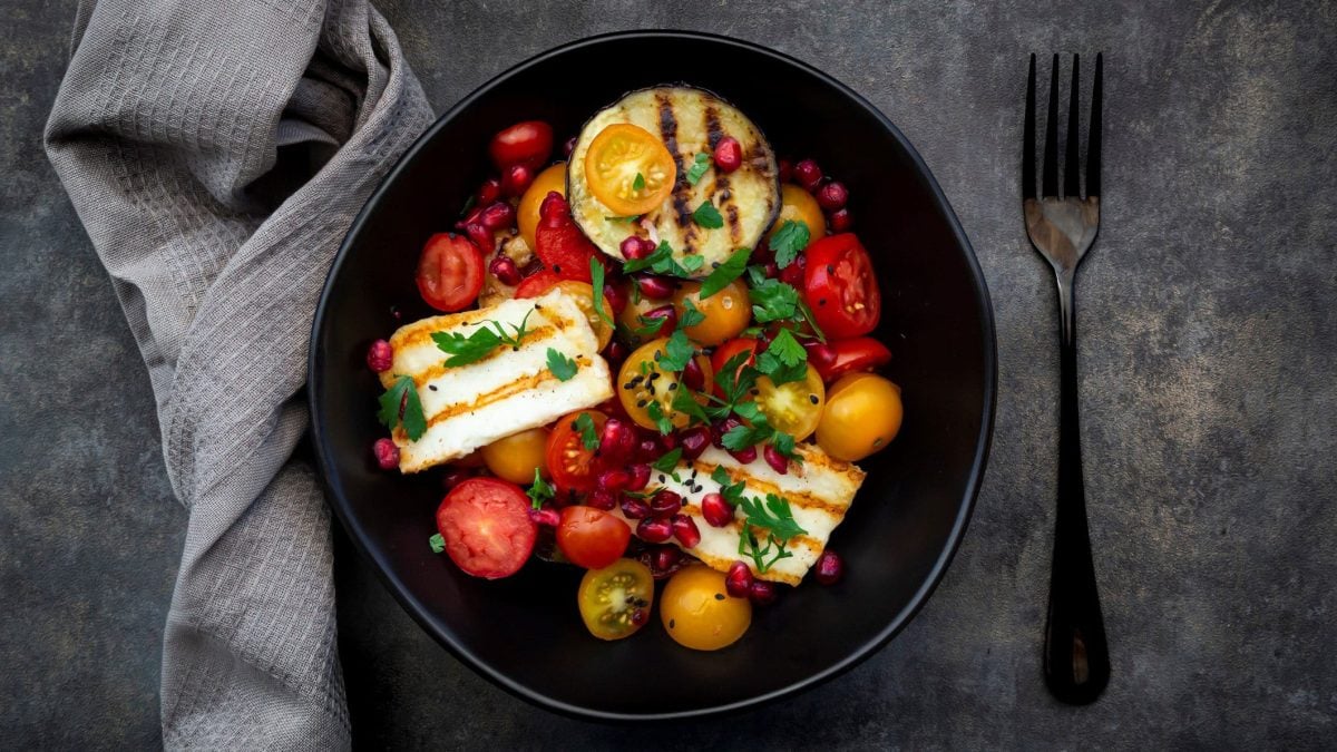 Eine schwarze Schale mit dem Tomatensalat und gegrillter Aubergine und Halloumi auf grauem Untergrund. Daneben ein graues Tuch und eine schwarze Gabel.