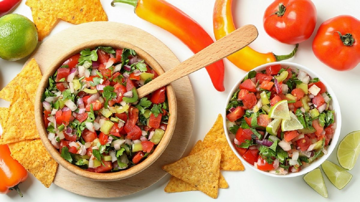 Zwei Schalen Pico de gallo, einmal Holz, einmal weißes Porzellan, auf hellem Untergrund. Tomaten, Chilis, Limette und Nachos als Dekoration im Bild. Aufnahme aus der Vogelperspektive.
