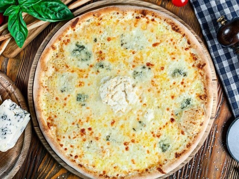 Auf einem Brettchen aus Holz liegt eine Gorgonzola-Pizza mit Rucola. Drumherum liegen Gorgonzola, TOmates, eine Pfeffermühl, Grisini und Basilikumblätter.