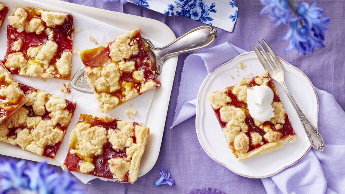 Prasselkuchen auf einer Servierplatte und auf einem Teller. Außerdem blaue Blumen. Das Ganze auf einer lilafarbenen Tischdecke.