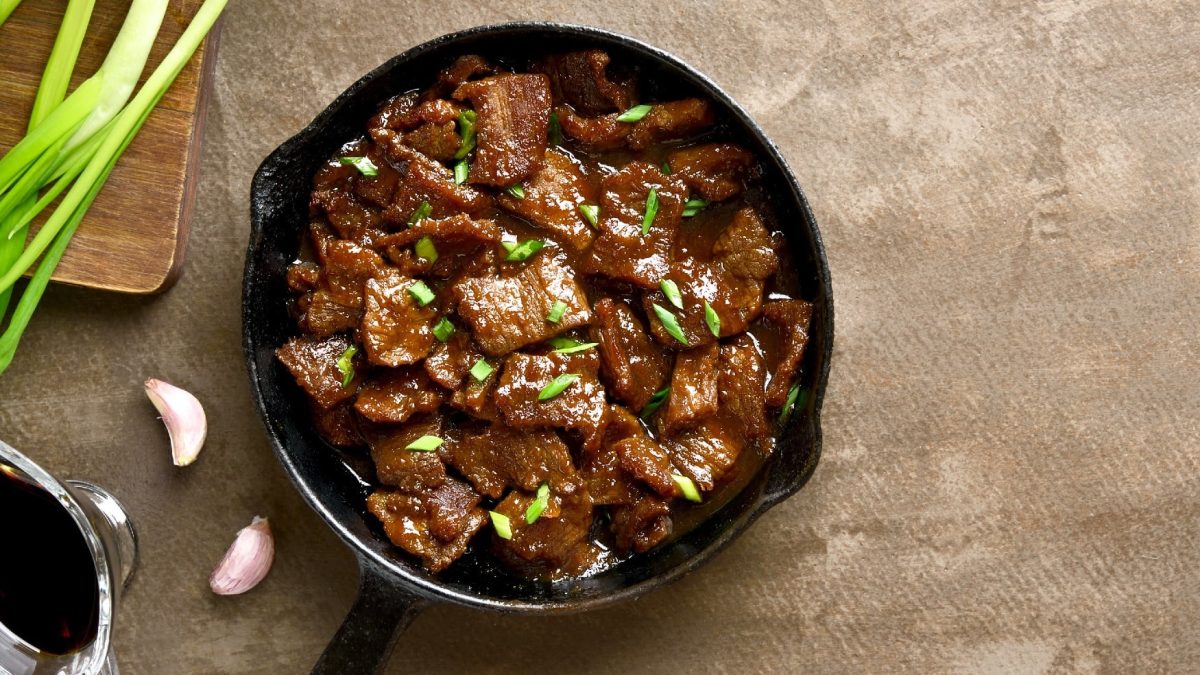 Eine schwarze Pfanne mit Abgoosht Yemen jemenitischer Rindfleischeintopf auf beigem Untergrund. Links davon Frühlingszwiebel und Knoblauch auf einem Holzschneidebrett.