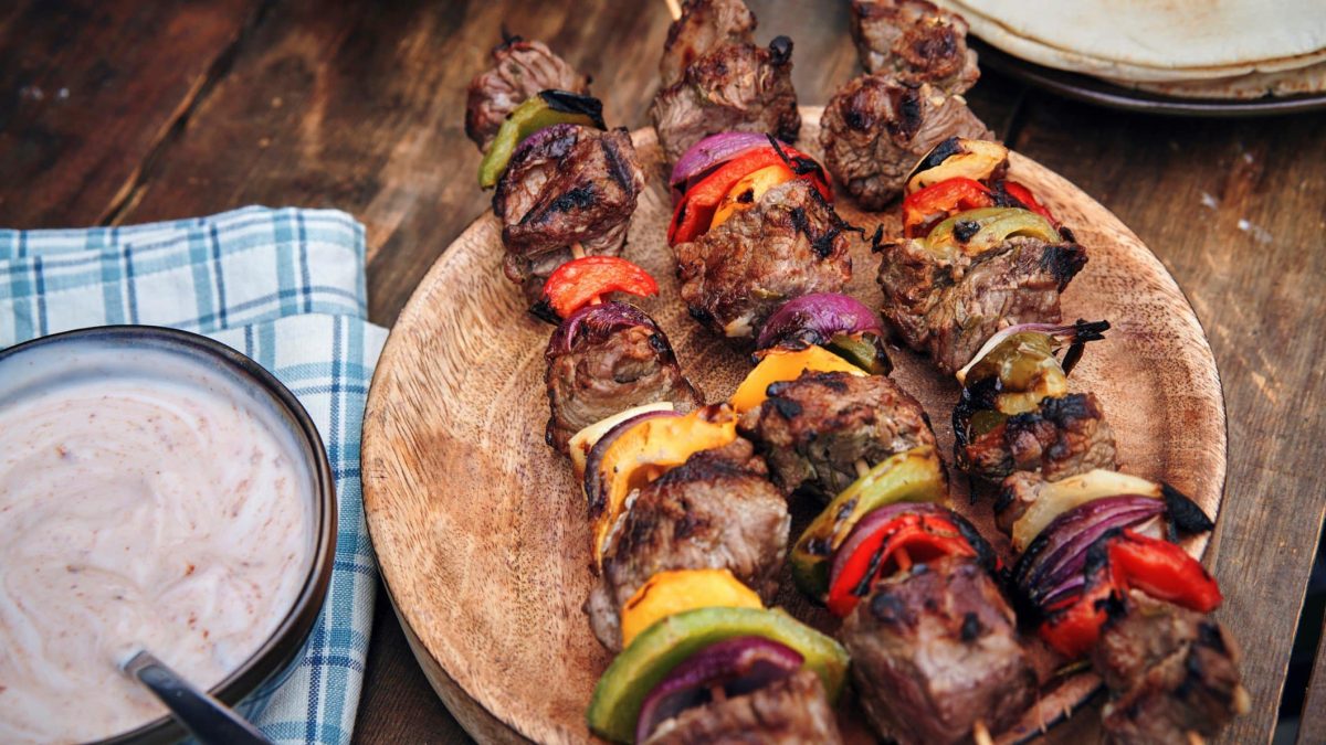 Türkische Shish Kebab mit Tomaten, Zwiebel und Paprika türkische Lammspieße auf rundem Holzbrett auf Holztisch. Daneben Schale mit hellem Dip auf blau-weißem Tuch. Im Hintergrund Fladenbrot. Aufnahme aus der Vogelperspektive.