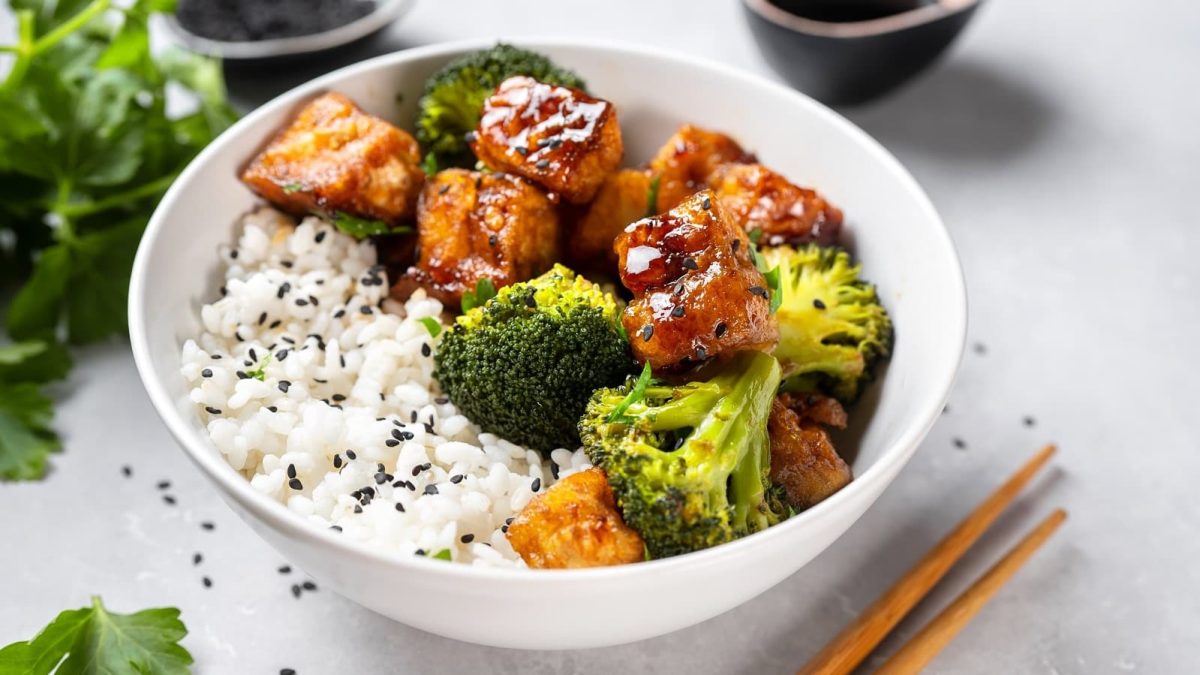 Tofu in Pfeffer-Sojasauce mit Brokkoli in einer Schüssel mit Reis neben einem Paar Stäbchen
