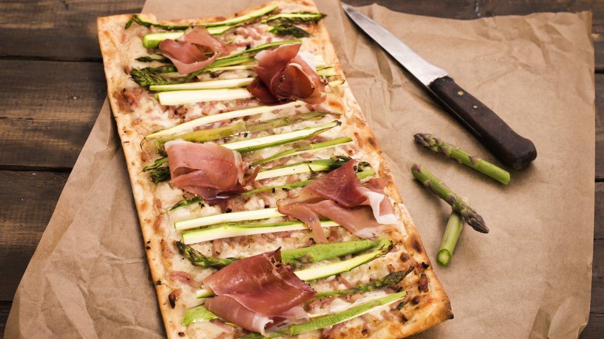 Spargel-Flammkuchen mit Serranoschinken auf einem Backpapier. Daneben etwas Spargel und ein Messer.