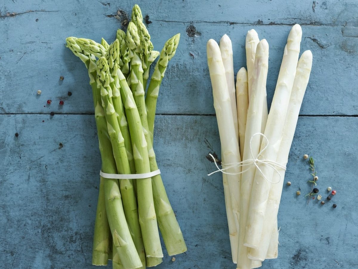 Gebündelter grüner und weißer Spargel auf einer hellblauen Platte mit der Anleitung, wie man Spargel richtig schälen kann.