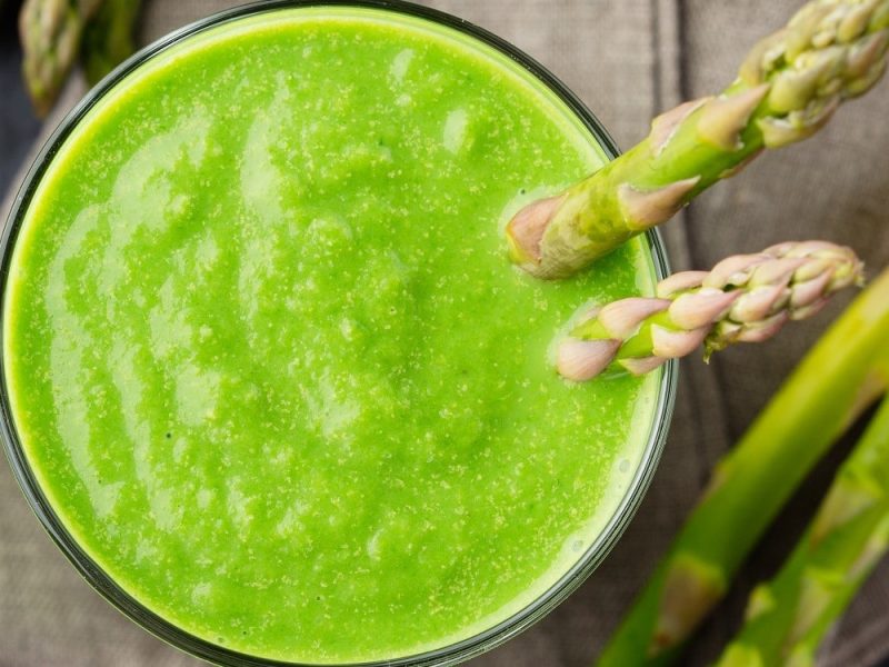 Grüner Spargel-Smoothie in einem Glas, garniert mit zwei grünen Spargel-Stangen auf einem hölzernen Tisch.