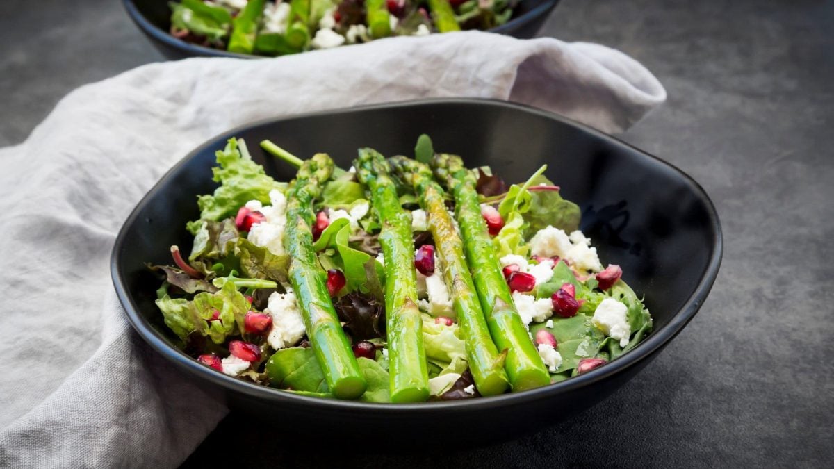 Gemischter grüner Salat mit Feta, Granatapfelkernen und gebratenem grünen Spargel in einer schwarzen Schale auf dunklem Untergrund.