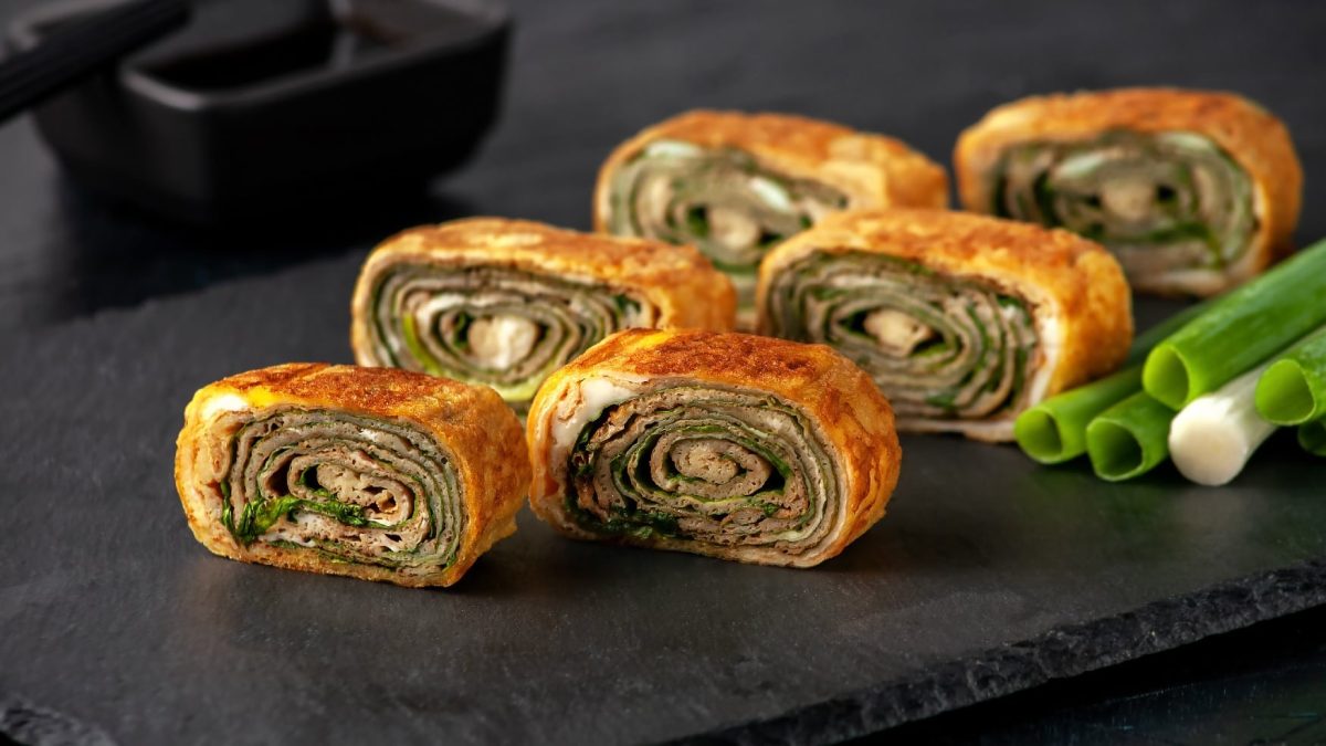 Tamagoyaki mit Pilzen und Lauch in 6 Stücke geschnitten auf einer schwarzen Schieferplatte. Im Hintergrund ein schwarzes Schälchen mit Sojasauce und Essstäbchen.