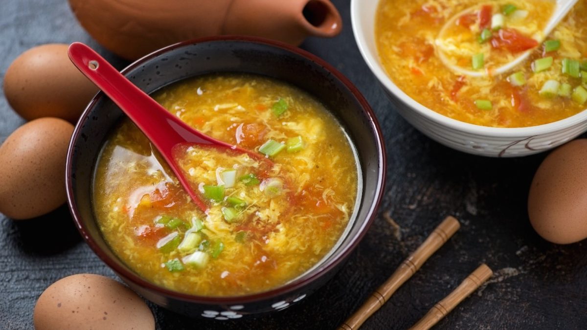 Egg-Drop-Suppe mit Tomate in einer schwarzen Schüssel auf einer schwarzen Steinoberfläche neben ein paar Eier und Stäbchen