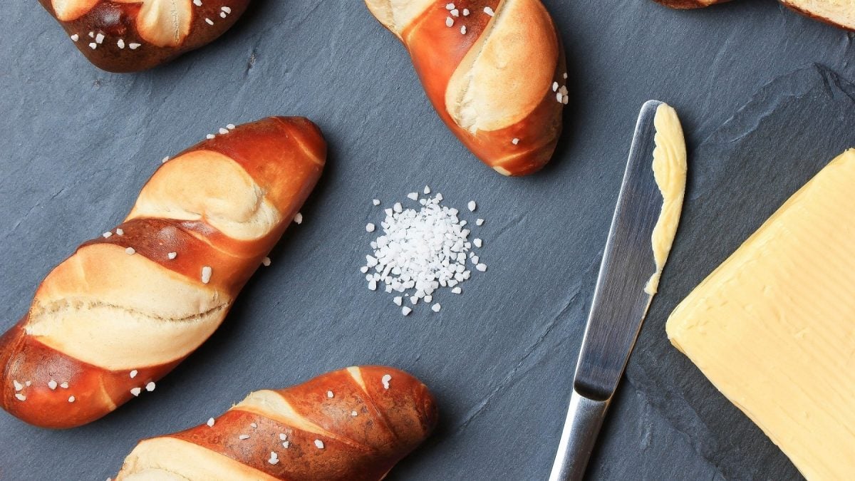 Vegane Laugenstangen, ein Buttermesser, Butter und Salz auf grauem Untergrund in der Draufsicht.