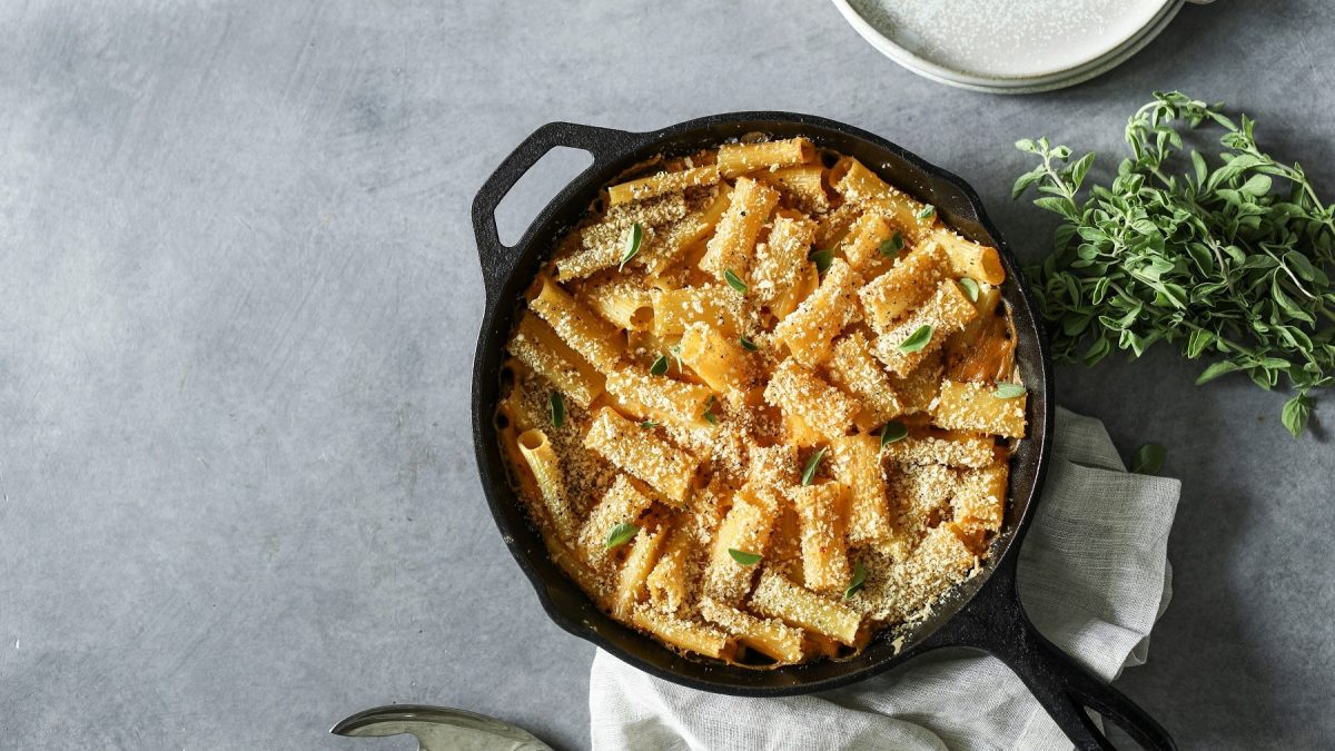 Eine gusseiserne Pfanne vegane Mac'n'Cheese mit Kräutern bestreut in der Draufsicht, daneben liegt ein Kräuterbund.
