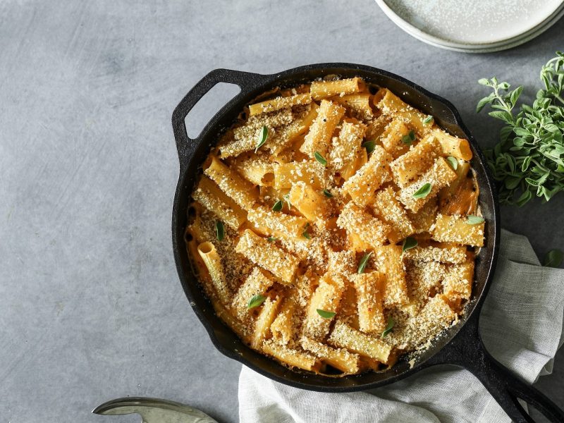 Eine gusseiserne Pfanne vegane Mac'n'Cheese mit Kräutern bestreut in der Draufsicht, daneben liegt ein Kräuterbund.