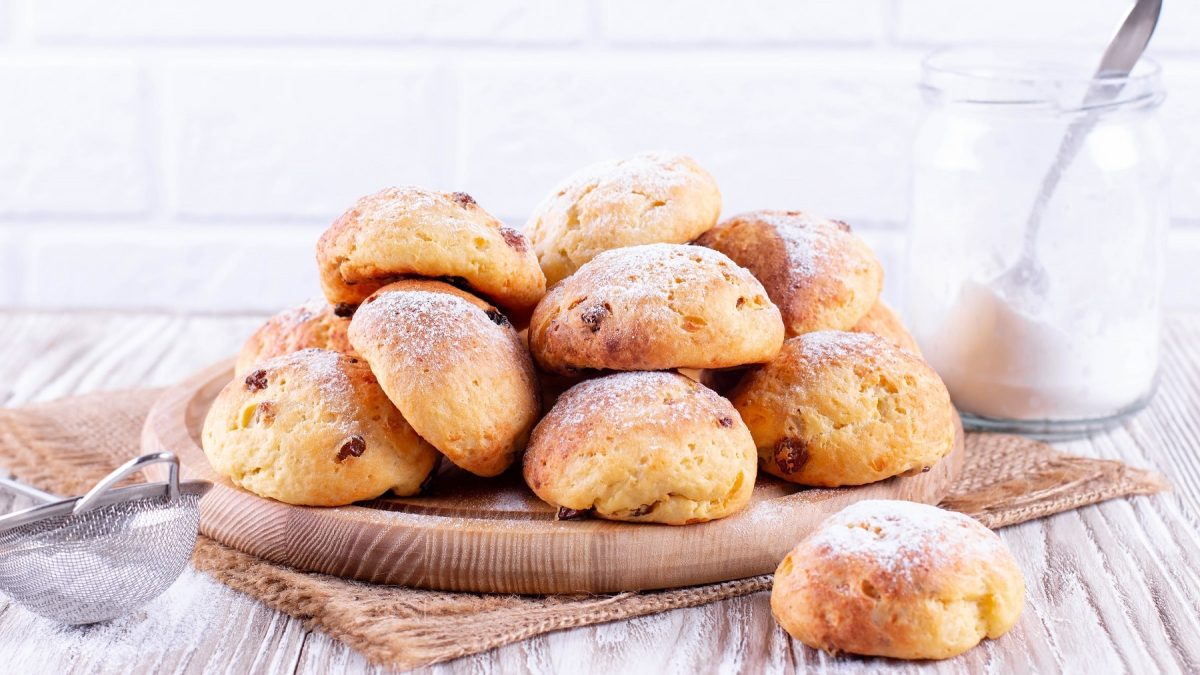 Ein Haufen veganer Rosinenbrötchen auf einem runden Holzbrett in hellem Setting, daneben ein Sieb und ein Glas Puderzucker.