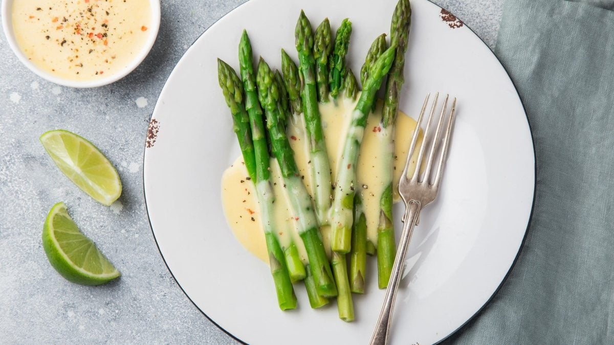 Ein Teller grüner Spargel mit veganer Sauce hollandaise und einer Gabel in der Draufsicht, daneben Limettenschnitze und eine Schale der Sauce.
