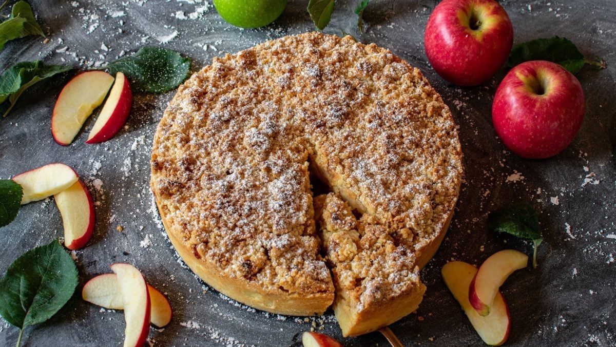 Auf einem Kuchenteller liegt ein veganer Apfel-Marzipan-Kuchen. Er ist mit Streuseln und Puderzucker bedeckt.
