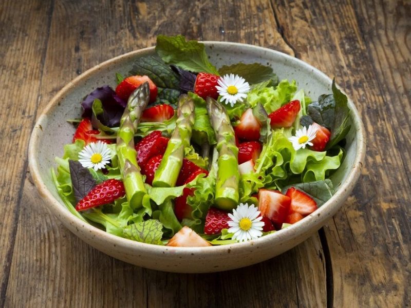 Auf einem hölzernen Untergrund steht eine große Schüssel. In der chüssel ist ein Wildkräutersalat mit grünem Spargel und Erdbeeren.