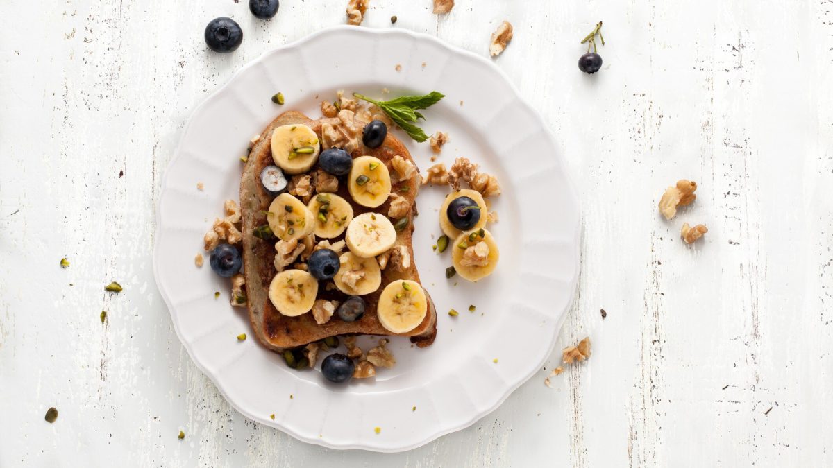 Arme Ritter mit Banane, Blaubeeren und Walnuss auf weißem Teller.