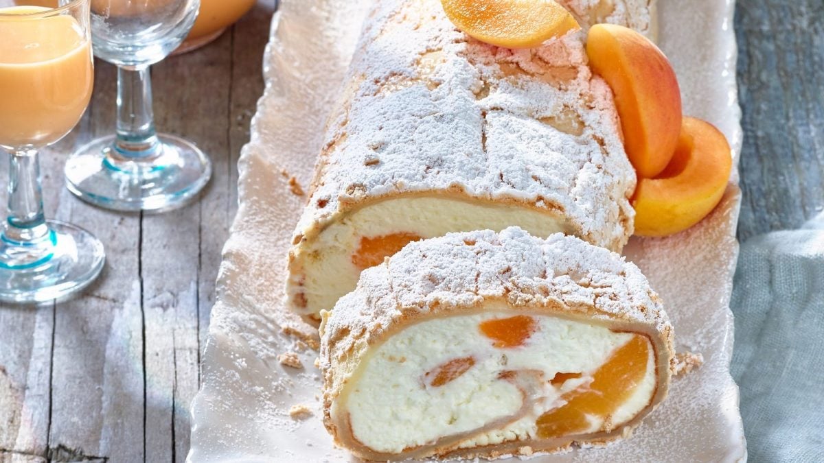 Auf einer Platte liegt eine Aprikosen-Pavlova-Rolle. Sie ist angeschnitten. Man erkennt die Füllung und die Aprikosen.