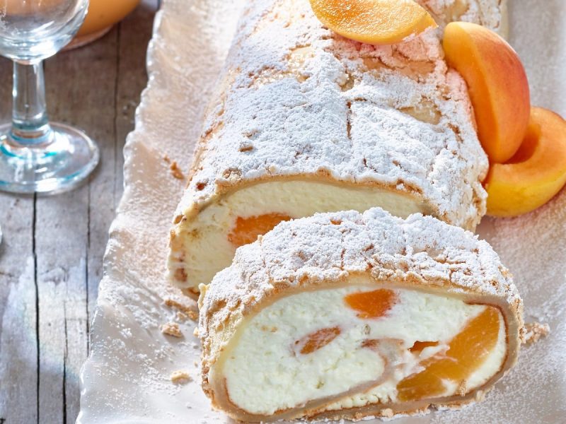 Auf einer Platte liegt eine Aprikosen-Pavlova-Rolle. Sie ist angeschnitten. Man erkennt die Füllung und die Aprikosen.