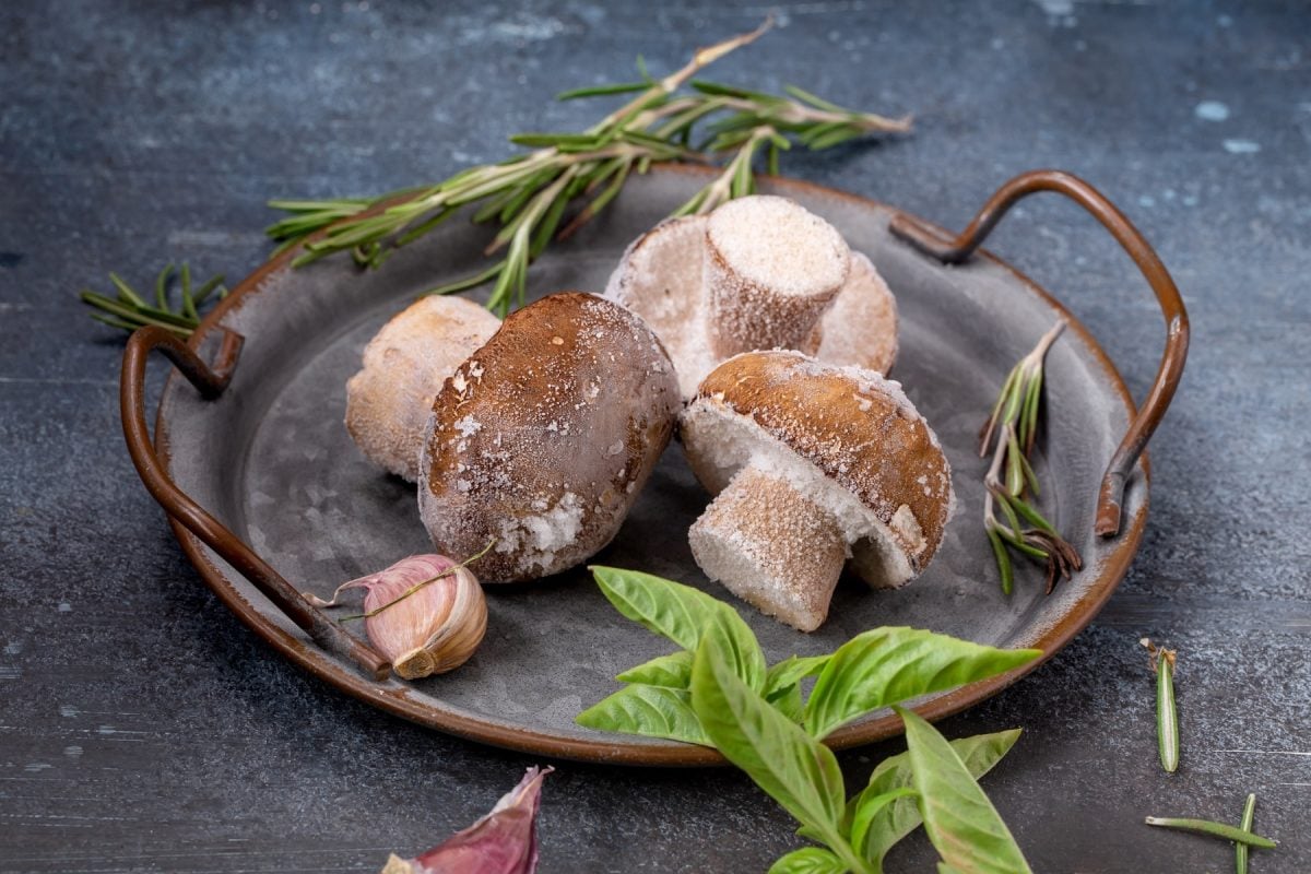 Drei gefrorene Pilze nebst Knoblauch und Kräutern in einer Metallschale.