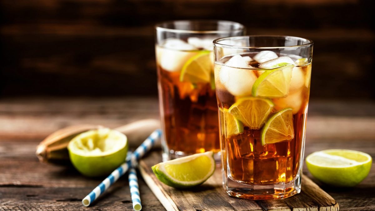 Zwei Gläser Long Island Iced Tea auf hölzernem Untergrund. Daneben liegen angeschnittene Limetten, ein Stößel und Strohhalme.