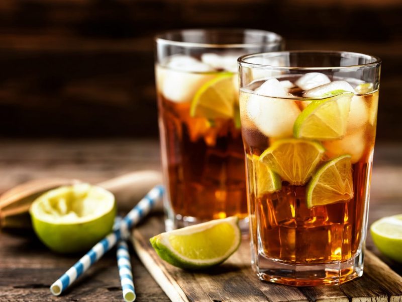 Zwei Gläser Long Island Iced Tea auf hölzernem Untergrund. Daneben liegen angeschnittene Limetten, ein Stößel und Strohhalme.