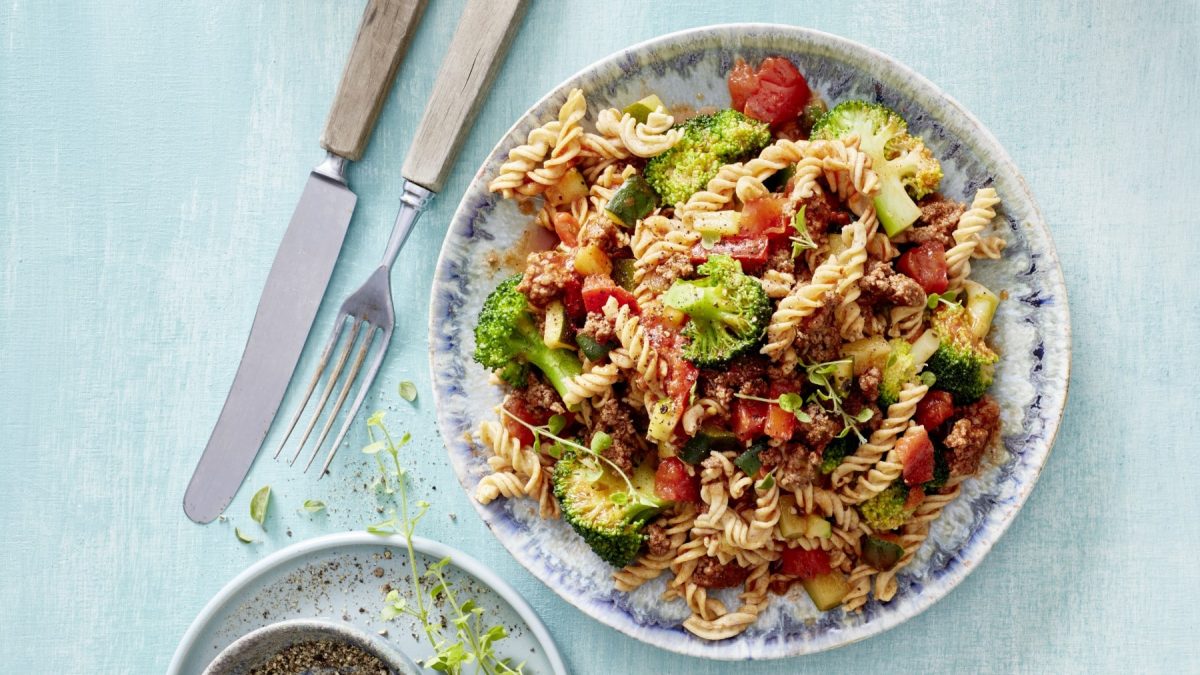 Nudelsalat mit Hack und Gemüse auf einem gemusterten Teller. Daneben Gewürze und Besteck.