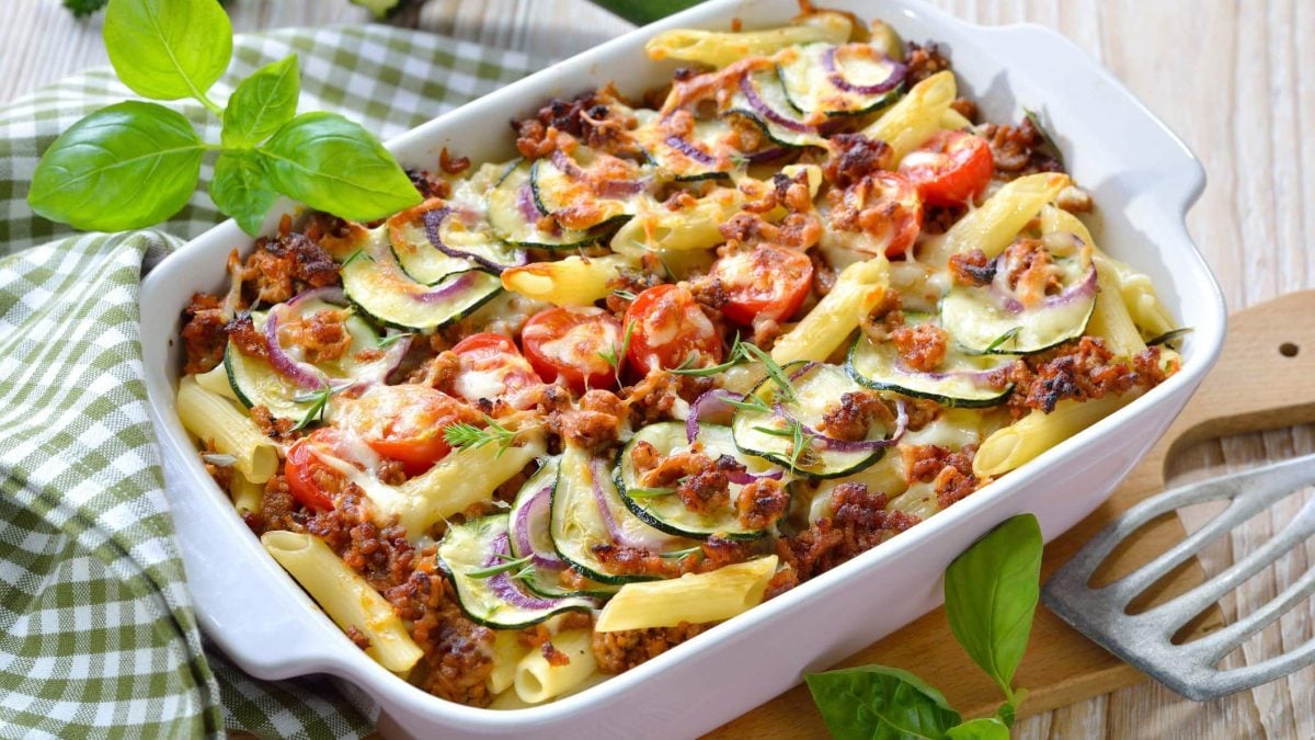 Pasta al forno mit Hackfleisch und Zucchini in weißer Auflaufform. Daneben liegt ein weiß-grün-kariertes Tischtuch