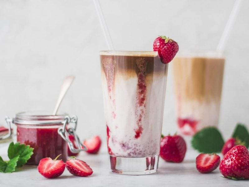 Ein Glas Iced Flat White mit Erdbeermilch und Erdbeersaice neben frischen Erdbeeren und einem Einmachglas