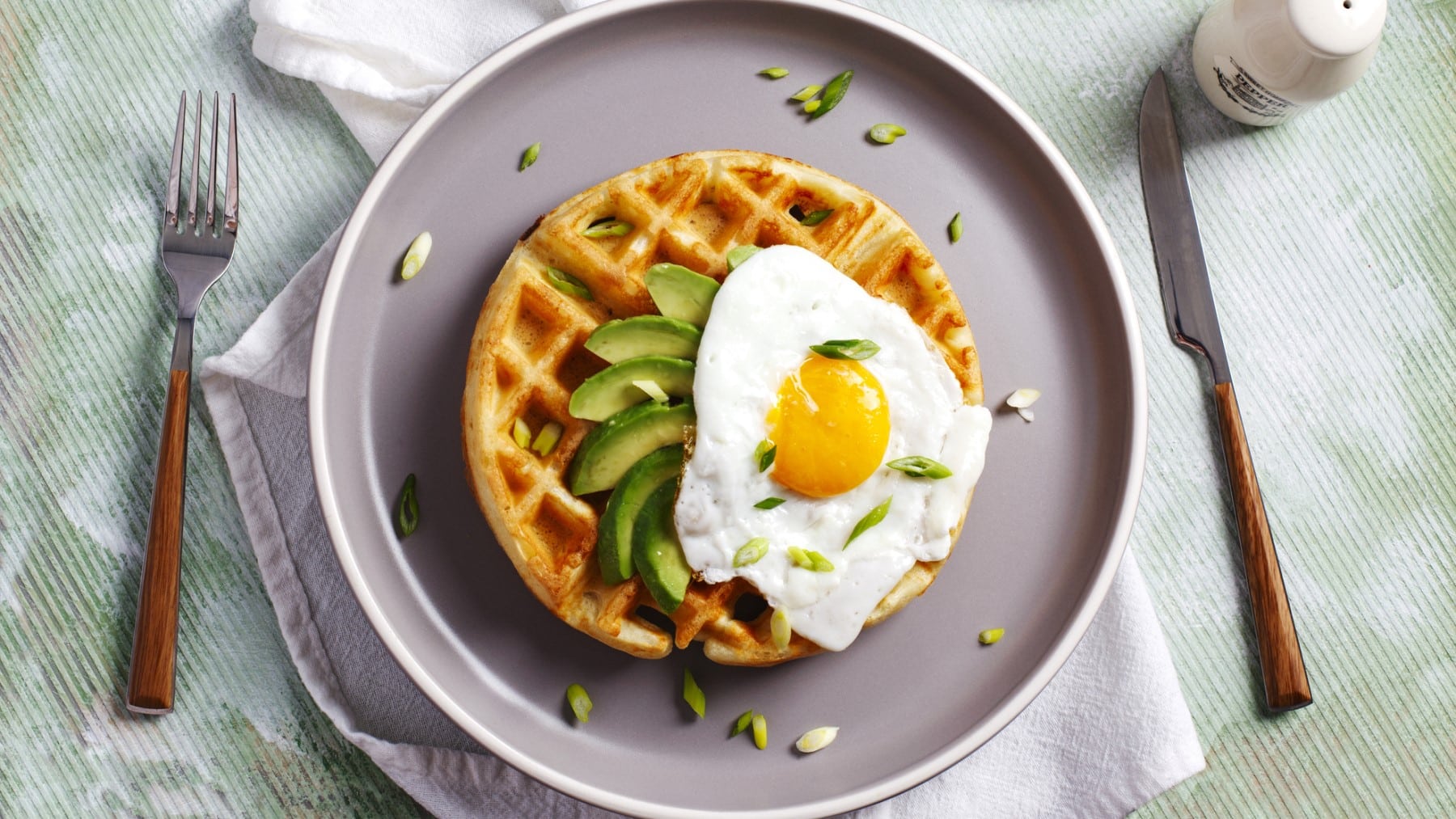 Waffel mit Avocado und Spiegelei auf einem grauen Teller. Daneben Besteck und ein Salzstreuer.