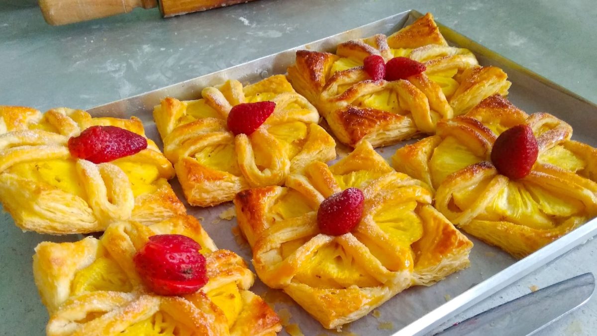 Sechs Ananas-Erdbeer Puffs auf einem Backblech mit Backpapier auf einem steinuntergrund und einem Küchenmesser.
