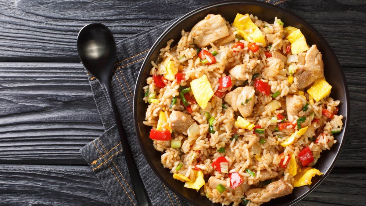 Arroz Chaufa peruanischer Reis mit Huhn in schwarzer Gusspfanne auf Jeanstuch und dunklem Holz. Daneben schwarzer Löffel. Aufnahme aus der Vogelperspektive.