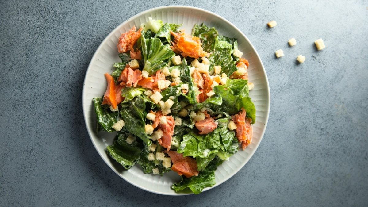 Caesar Salat mit Lachs aus einem weißen Teller. Daneben liegen Croutons.