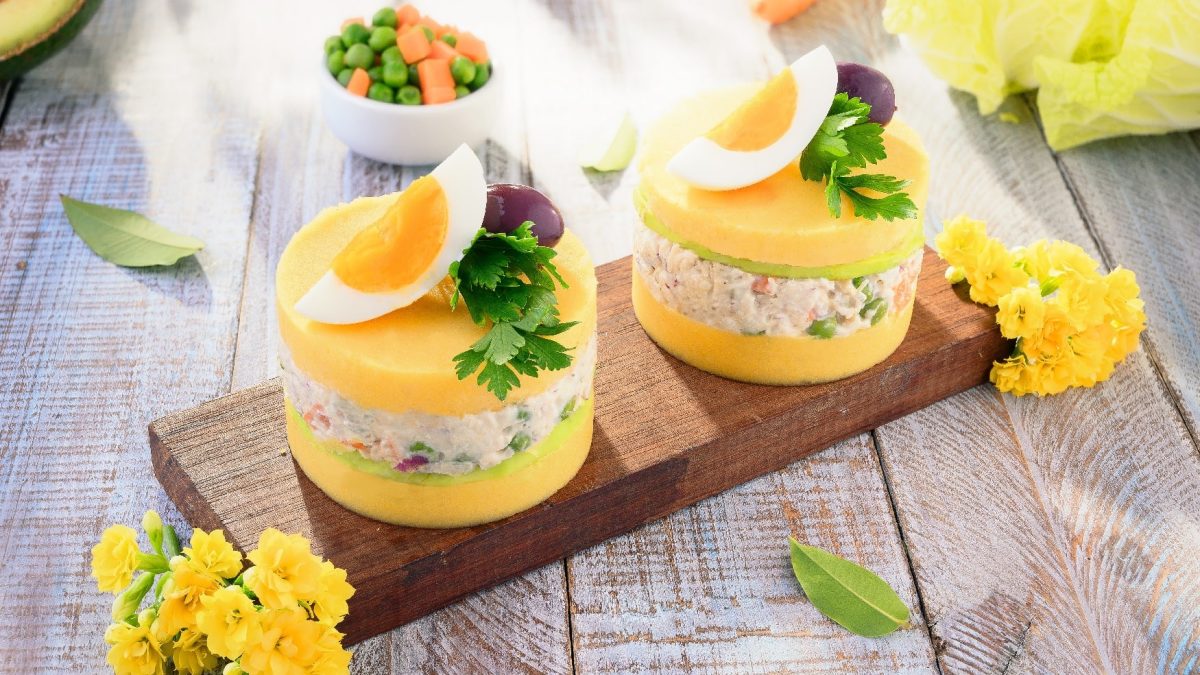 Zwei Portionen Causa limena auf einem Holzbrett mit Blüten und Gemüse drum herum auf einem Holztisch.