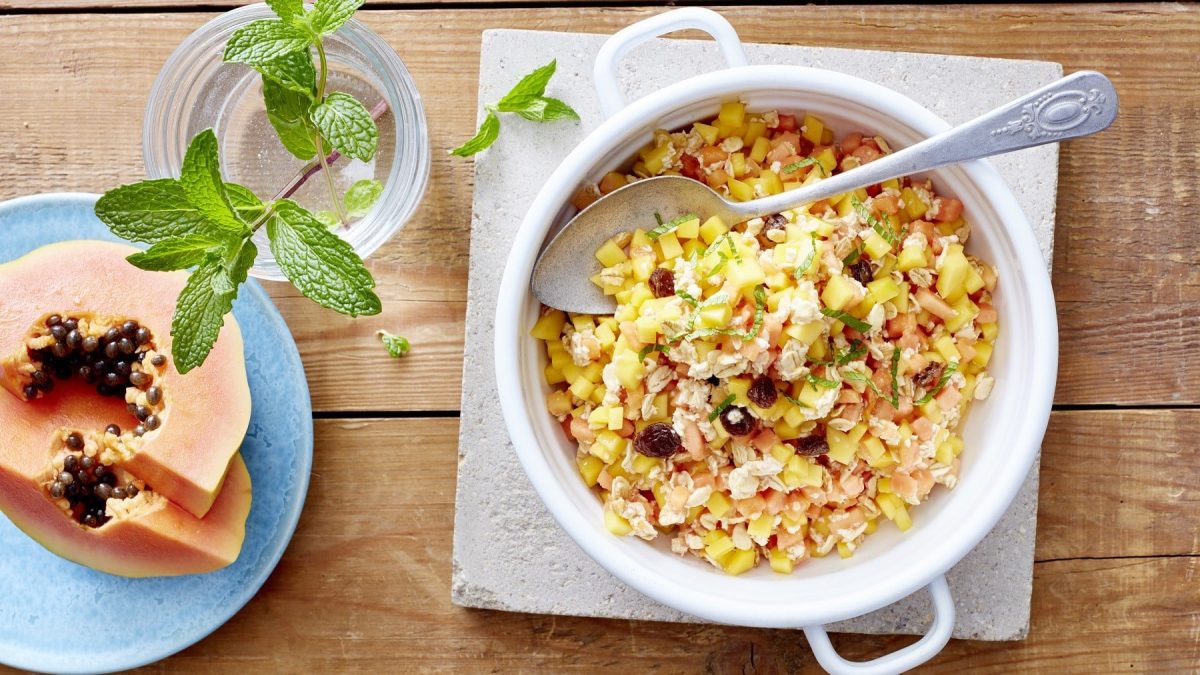 Exotisches Müsli mit Mango, Papaya, Nüssen, Rosinen und Kokosflocken, garniert mit frischer Pfefferminze und serviert in einer weißen Schale auf einem klassischen Holztisch