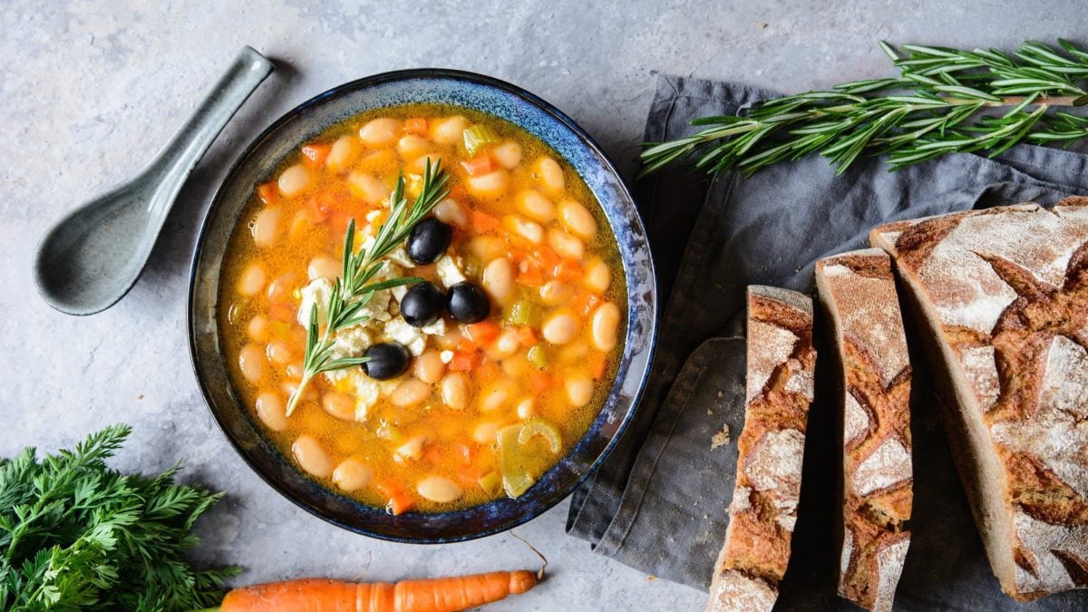 Eine dunkelblaue Suppenschale mit der Bohnensuppe auf grauem Tisch mit Bundmöhren und Landbrot an der Seite.