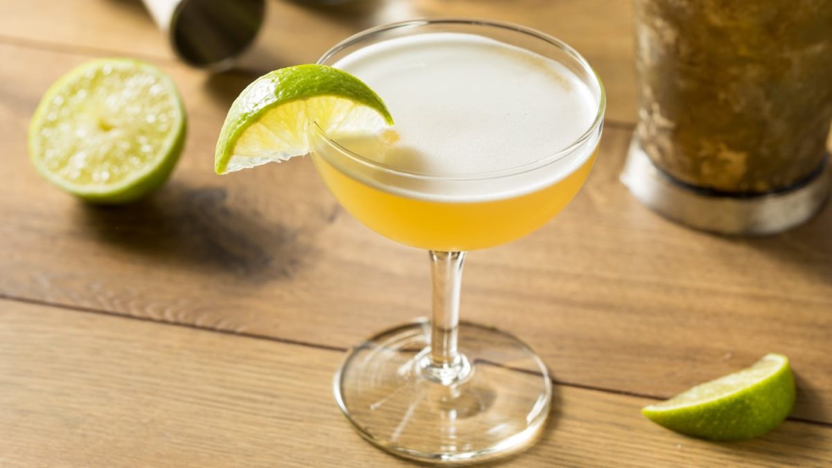 Ein Glas mit Gimlet auf einem Holztisch mit Limetten und Bar-Equipment.