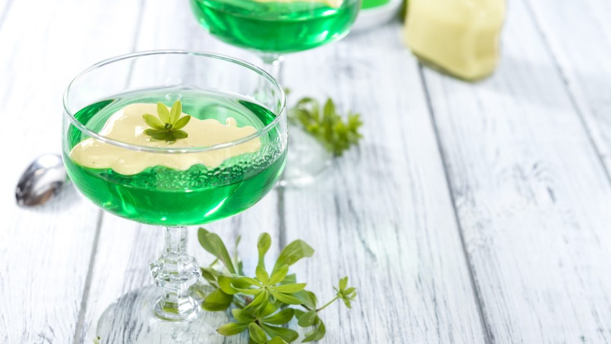 Zwei Gläser mit grüner Götterspeise und Vanillesauce auf einem weißen Holztisch mit frischem Waldmeister.