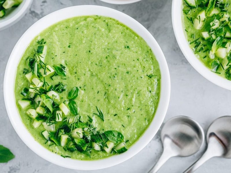 Zwei Schalen mit einen Gurken-Gazpacho steht auf einem Tisch. Daneben liegen zwei Löffel.