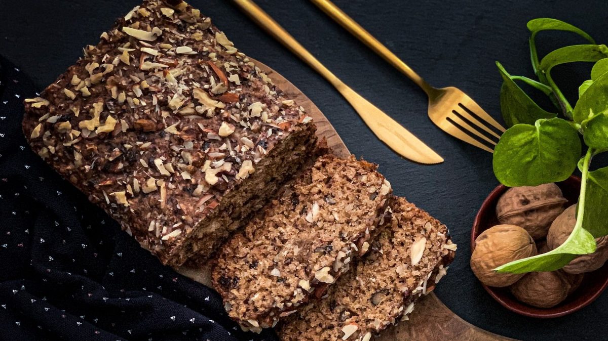 Ein angeschnittenes Haferflocken-Brot ohne Backen in der Draufsicht, daneben liegt goldenen Besteck, außerdem sind Walnüsse in einer Schale zu sehen.