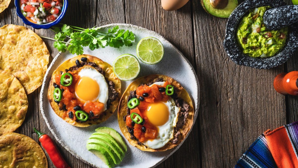 Huevos ranchero mexikanisches Frühstück auf Teller mit Limette und Avocado. Daneben Tortilla, Salsa und Guacamole. Chilis, Avocados und Tomaten als Dekoration im Bild. Arrangiert auf Holztisch. Aufnahme aus der Vogelperspektive.