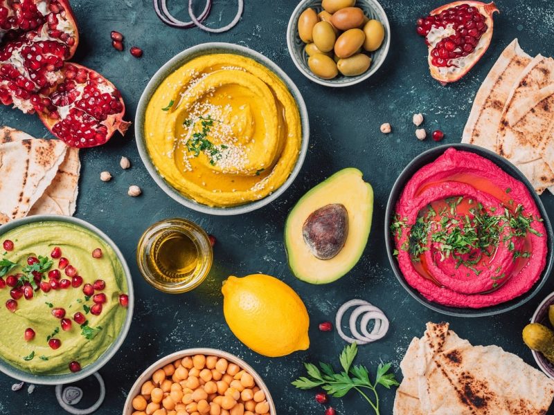 Mehrere Schalen mit Hummus stehen auf einem dunklem Hintergrund. Dazu auch noch eine halbe Avocado, eine Zitrone und Granatapfel.