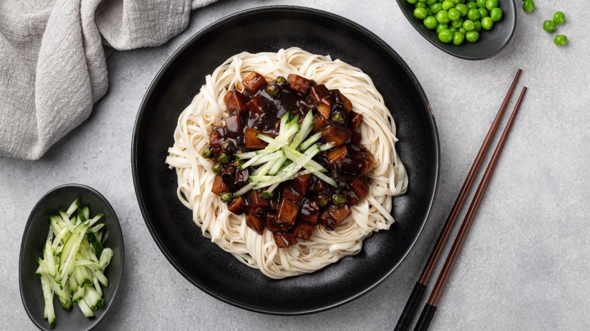 Eine Schüssel Jajangmyeon auf einem grauen Tisch neben einer Schale Gurkenstreifen und Erbsen