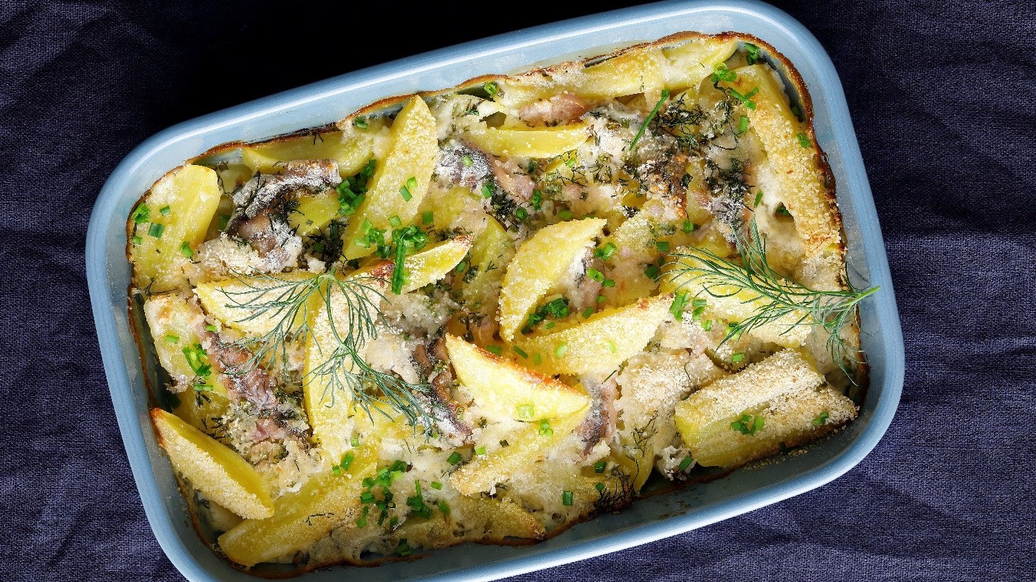 Eine Auflaufform mit Kartoffel-Anchovi-Auflauf auf einem dunklen blauen Tuch.